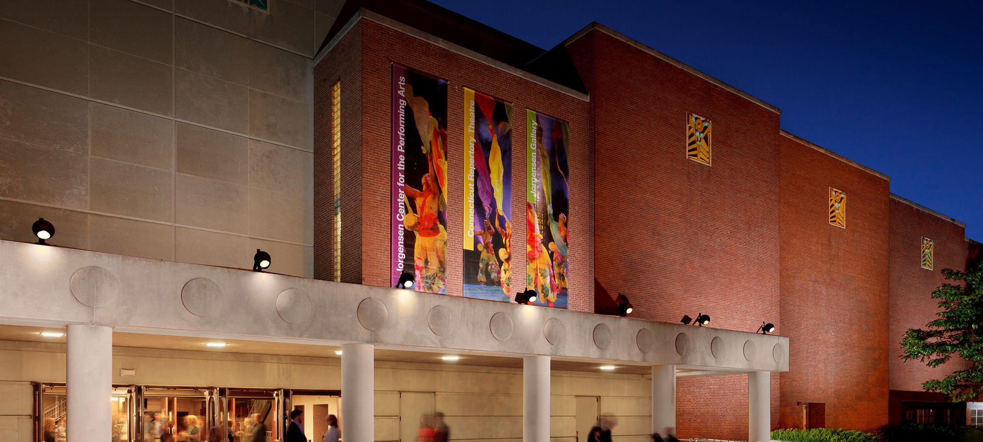 Exterior view of the Jorgensen Center at dusk, featuring a brick and concrete facade with colorful vertical banners and illuminated entrance doors where people are entering.