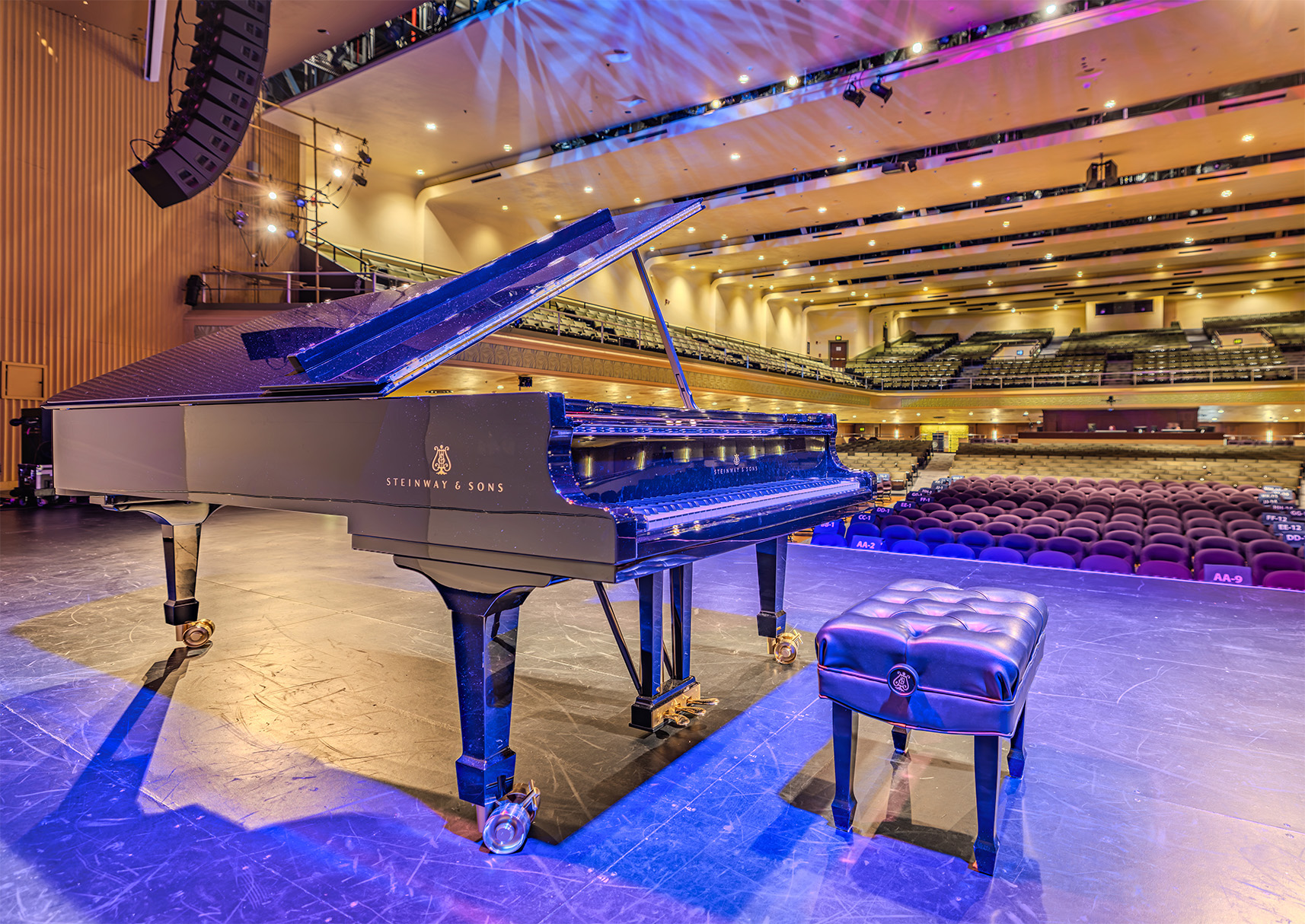 A Steinway & Sons grand piano sits on stage under warm lighting in an empty concert hall, with rows of seats visible in the background.