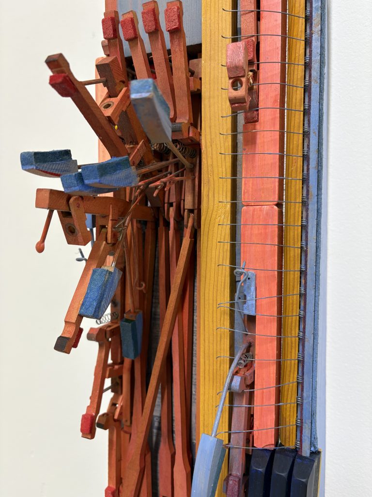 Close-up detail of a colorful wooden assemblage featuring layered rods, springs, and painted blocks arranged in a vertical sculptural composition